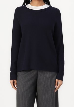 Eine Frau trägt einen dunkelblauen Pullover über einem weißen Hemd, kombiniert mit grauen, gestreiften, weit geschnittenen Hosen, und steht vor einem weißen Hintergrund.