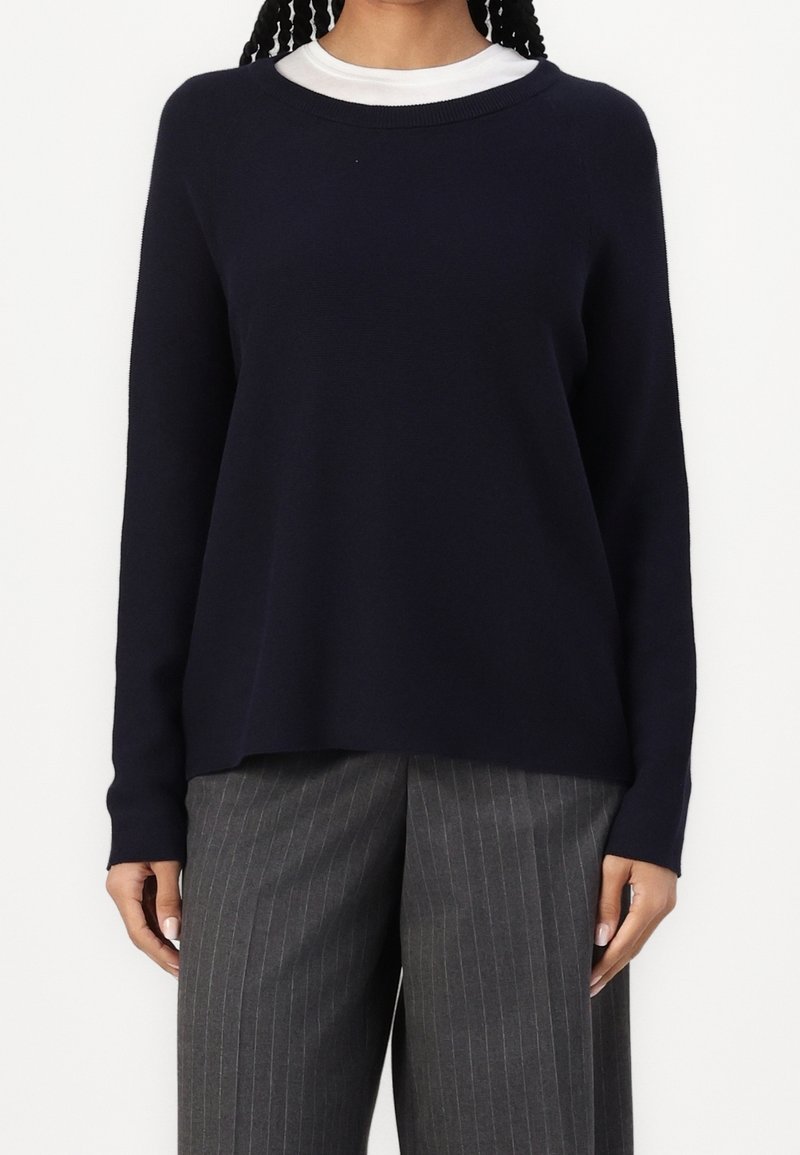 Eine Frau trägt einen dunkelblauen Pullover über einem weißen Hemd, kombiniert mit grauen, gestreiften, weit geschnittenen Hosen, und steht vor einem weißen Hintergrund.