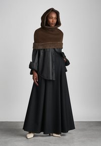 Copertura marrone con cappuccio e rifinitura in pelliccia, indossata sopra una camicia in pelle nera, abbinata a una gonna nera fluida e scarpe chiare.