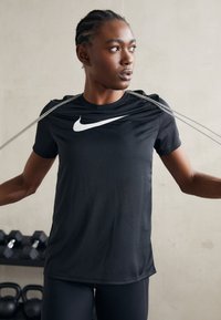 Črna majica s kratkimi rokavi Nike z belim logotipom, gladka tekstura in prilegajoča zasnova, jo nosi oseba, ki drži upogibne elastike.