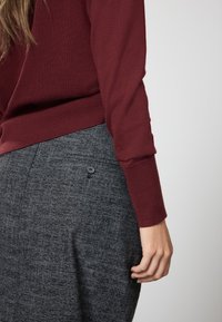 Bordo spalvos megztinis ilgomis rankovėmis derinamas su pilku tekstūruotu sijonu, kuriame yra šoninė kišenė su juoda sagos detalė.