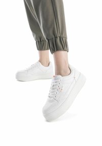 Zapatillas blancas con una parte superior de cuero suave, con acentos sutiles, una suela texturizada y un diseño de plataforma gruesa, combinadas con pantalones verdes doblados.
