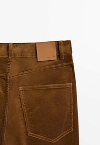 Pantalon en velours côtelé marron montrant la poche arrière et un patch de marque en cuir fauve sur la ceinture.