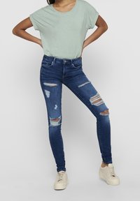 Ljusgrön kortärmad skjorta i kombination med blå tajta jeans med flera slitna områden och hål, kompletterat med ljusa sneakers.