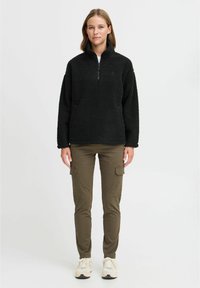 Black fleece pullover with a high collar and half-zip, paired with olive green cargo pants featuring side pockets, worn with light sneakers.