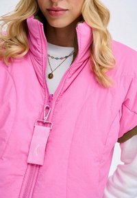 Růžová péřová vesta s vysokým límcem a zapínáním na zip. Má strukturovaný povrch, nastavitelný popruh a kontrastní zelenou podšívku.