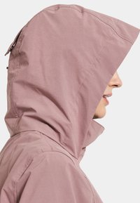 Ljusrosa vattentät jacka med huva, en strukturerad huva, en slät yta och synlig sömnad längs sömmarna och halsringningen.