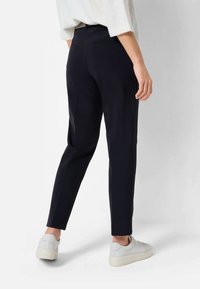 Pantaloni neri su misura con una vestibilità slim e una texture liscia, dotati di tasche posteriori e dettagli sui fianchi, abbinati a sneakers grigie chiare.