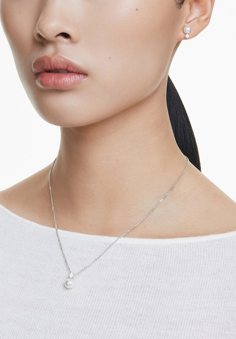 Hienoketjuinen hopeaketju, jossa on yksi pyöreä helmi-riipus, yhdistettynä matching helmi-korviksilla. Näkyvissä oleva teksturoidusta valkoisesta paidasta.