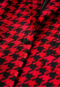 Rood en zwart houndstooth-patroonstof met een zachte textuur, voorzien van een zwarte knop en rafelige randen bij de kraag en naden.
