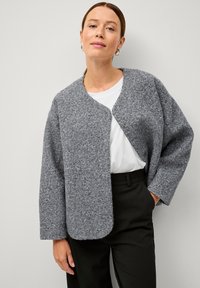 Giacca grigia in feltro con scollatura rotonda, apertura frontale e maniche ampie, indossata sopra una camicia bianca e pantaloni neri.