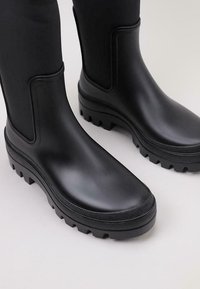 Bottines noires en caoutchouc avec une finition mate, dotées d'une semelle texturée, d'un bout rond et d'accents latéraux pour un enfilage facile.