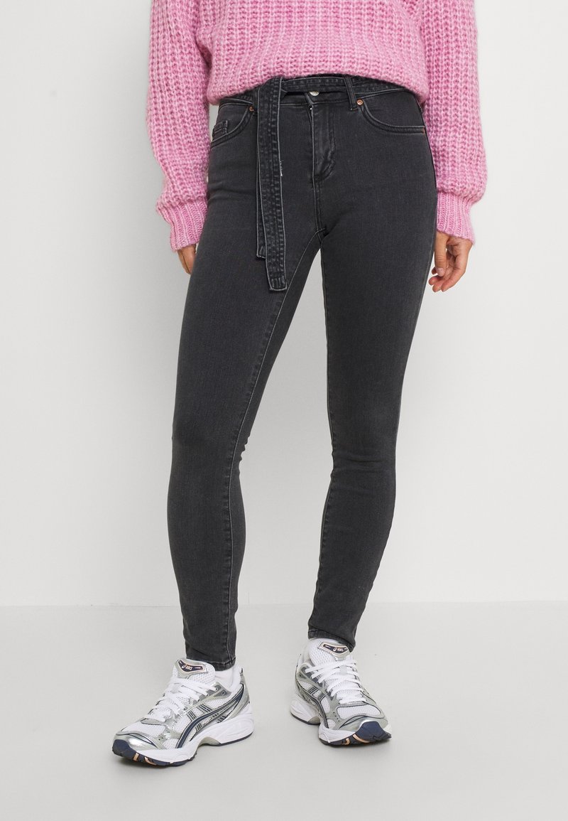 Jeans ajustados de tiro alto en color negro con cintura con cinturón, que presentan un diseño elegante y una textura de denim suave, combinados con zapatillas blancas y plateadas.
