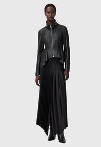 Schwarze Lederjacke mit hohem Kragen, durchgehendem Reißverschluss und Peplum-Saum; kombiniert mit einem schwarzen plissierten Maxirock und knielangen schwarzen Stiefeln.