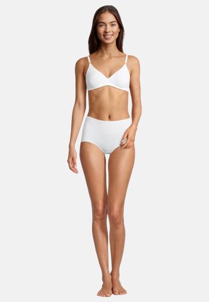 Woman standing barefoot, smiling, wearing white wireless bra and high-waisted briefs against plain light background.