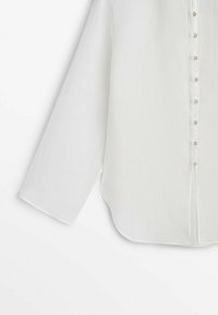Chemise blanche à manches longues avec boutons, ourlet cintré et boutons nacrés sur fond blanc uni.