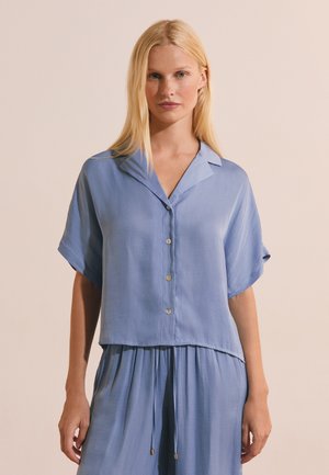 Femme blonde portant une chemise ample bleu clair à manches courtes et un pantalon à cordon assorti, debout devant un fond beige.