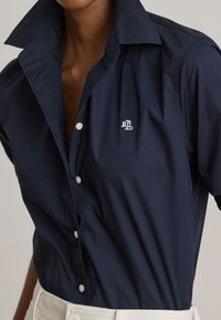 Asmuo dėvi tamsiai mėlyną marškinius su sagutėmis, pakelta apykakle ir baltu siuvinėtu „RL“ logotipu ant krūtinės, derinama su baltais kelnėmis.