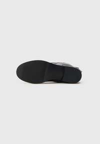Sola de bota de tornozelo em couro preto mostrada por baixo, com salto texturizado e antepé, num fundo branco liso.