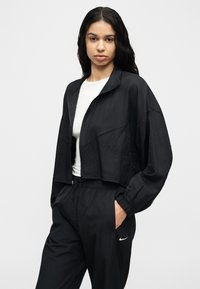 Νεαρή γυναίκα με μακριά σκούρα μαλλιά, φορώντας μαύρο μπουφάν με φερμουάρ πάνω από λευκό τοπ και μαύρα αθλητικά παντελόνια Nike, με τα χέρια στις τσέπες.