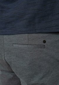 Graue, strukturierte Hose mit glattem Taschenbesatz und einem kleinen schwarzen Akzentknopf. Kombiniert mit einem dunkelblauen Stoffoberteil.