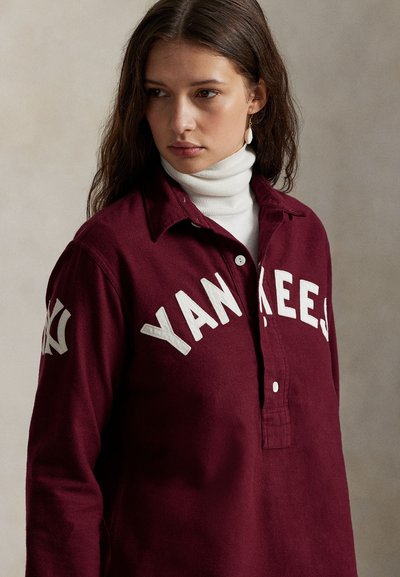 Μπορντό πουκάμισο με λευκό κείμενο "YANKEES", σχεδίαση με γιακά, κουμπάκια και μαλακή υφή υφάσματος, φορεμέν�ο πάνω από μια λευκή μπότα λαιμού.