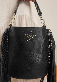 Schwarze Bucket-Bag aus genarbtem Leder mit Fransen an den Seiten, sternförmiger Naht und goldenen Sternnieten, gehalten von einer Hand in einem cremefarbenen Kleid.