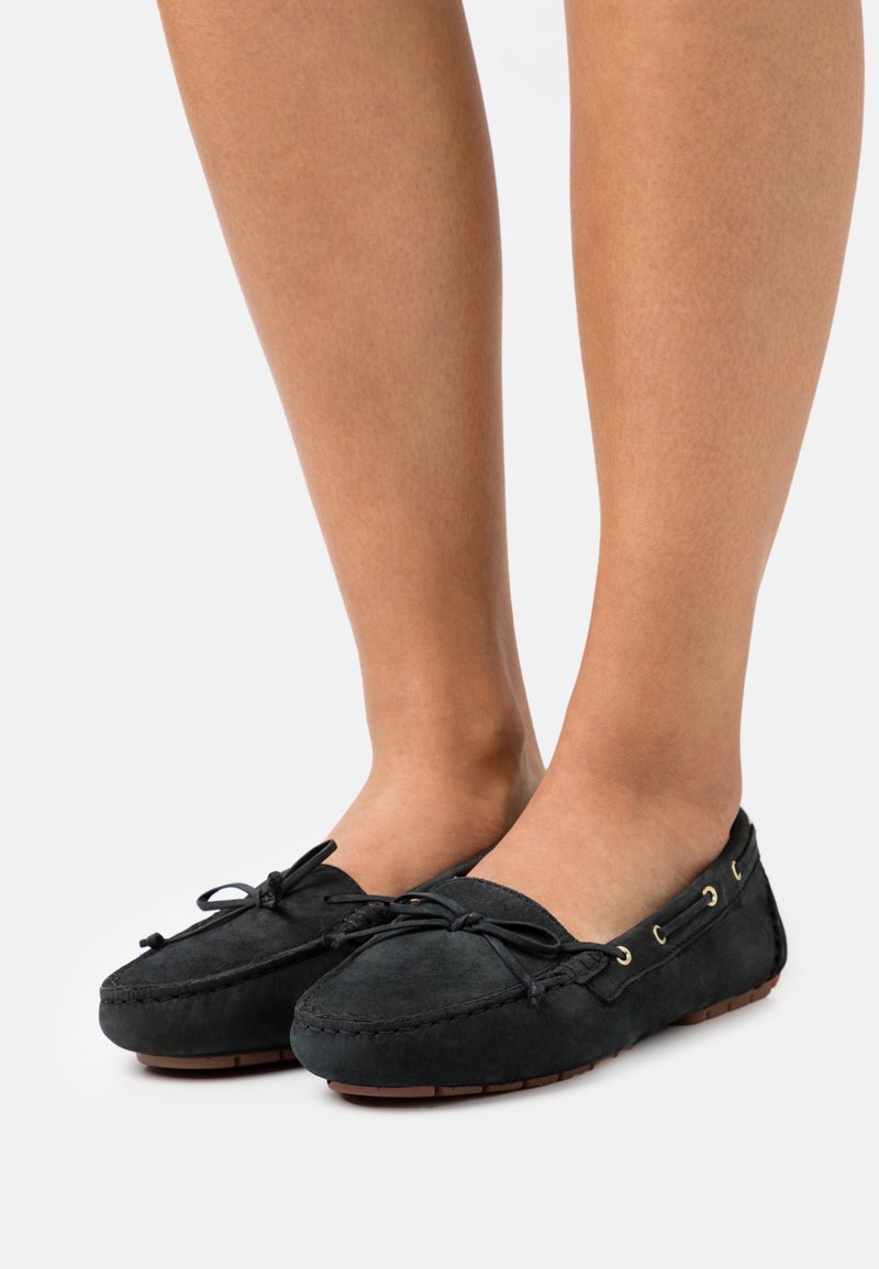 Chaussures mocassins en daim noir avec détails de nœud portées pieds nus, présentées sur un fond gris clair.