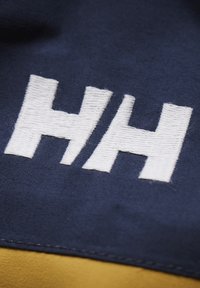 Námorní látka obsahuje vyšívané bílé logo "HH" s texturovaným pouzdrem, které zvýrazňuje kontrastní žlutý akcent pod ním.