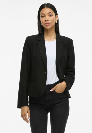 Sort blazer med en skræddersyet pasform, slagkrave, enkelt knaplukning og to frontlommer, båret over en hvid t-shirt og sorte jeans.