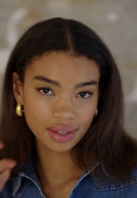 Nærbilde av en person med langt, mørkebrunt hår, iført gullhøye øreringer. Bakgrunnen er mykt uskarp. Huden har en naturlig glød.