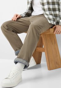 Khakifarbene Chinos mit aufgerollten Bündchen, innen mit einem blauen Karomuster gefüttert. Kombiniert mit weißen Turnschuhen auf einem Holzschemel.