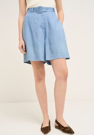Kvinde iført lyseblå shorts med høj talje og bælte, en hånd i lommen, kombineret med leopardmønstrede lavhælede slingback-sko.