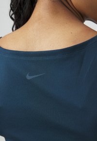 Tumedam sinine sporditop ribatekstiiliga, laia kaelusega ja seljaosas diskreetne Nike logo. Sile kangas ja vormiline disain.
