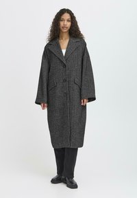 Kvinde med langt krøllet hår iført en lang mørkegrå herringbone-frakke, sorte bukser og sorte sko, som står foran en enkel hvid baggrund.