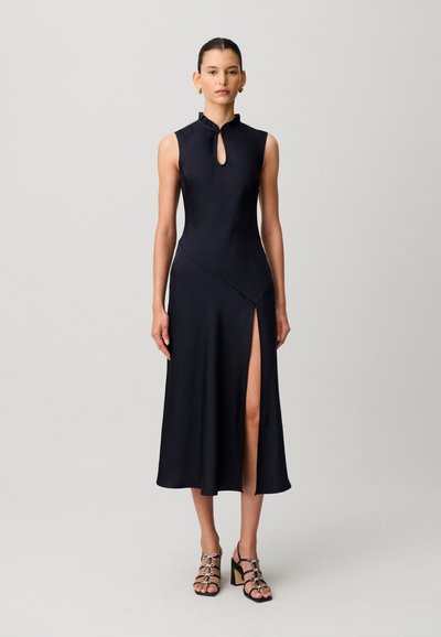 Mujer de pie vistiendo un vestido midi negro sin mangas con cuello alto, abertura en forma de ojo de cerradura, abertura lateral y sandalias negras de tacón bloque con tiras.