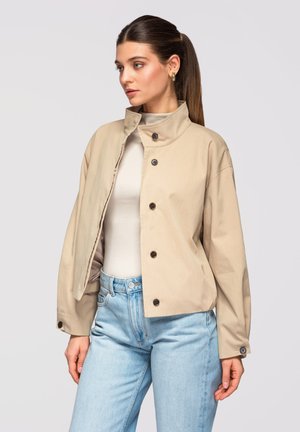 Frau mit braunen Haaren im Pferdeschwanz, die goldene Creolen trägt, eine beige Jacke mit Knöpfen über einem weißen Oberteil und hellblaue Jeans, blickt nach links.