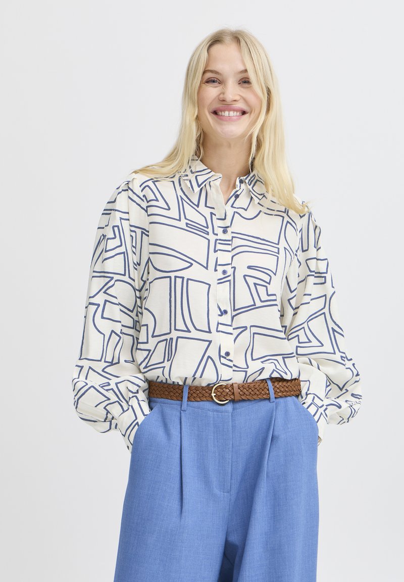 Camicia button-up color crema con motivo geometrico astratto blu, maniche lunghe, vestibilità rilassata; abbinata a pantaloni azzurri chiari e una cintura intrecciata marrone.