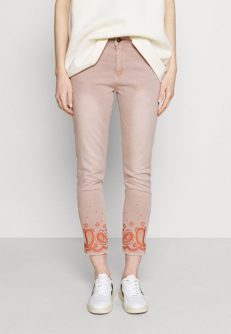 Jeans cortos rosas con intrincado bordado de paisley naranja en el dobladillo, hechos de denim. Estilizados con una blusa holgada clara y zapatillas blancas.