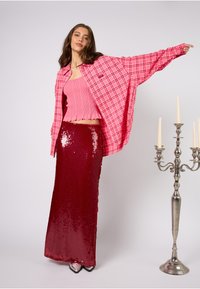 Różowa koszula w kratę w dużym rozmiarze, różowy prążkowany top odsłaniający brzuch oraz długa, czerwona spódnica z cekinami sięgająca do podłogi. Dodatkowe szczegóły: metaliczne buty i kandelabr.