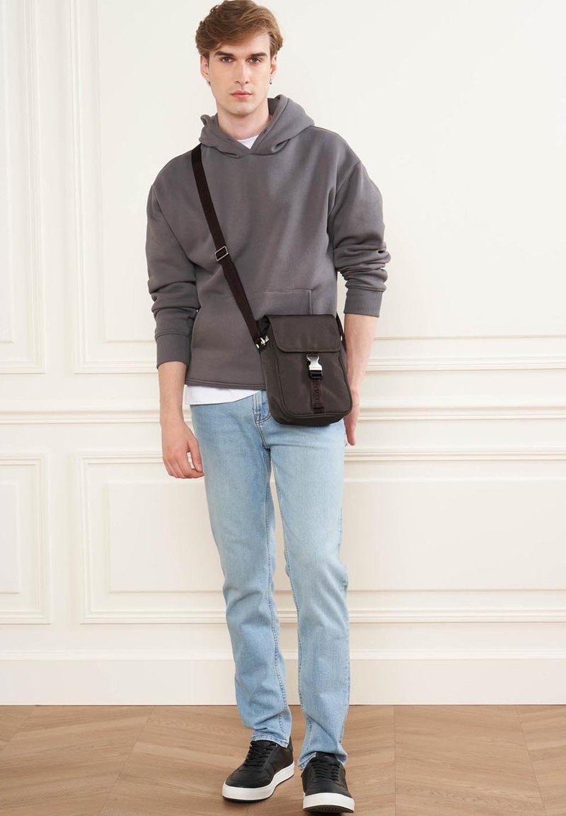 Hoodie grigia con tasca frontale, jeans azzurri chiari e sneakers nere. Borsa a tracolla marrone con pattina e dettaglio fibbia.