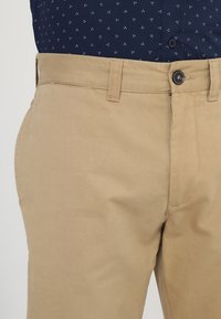 Pantaloni khaki con una texture liscia, dotati di chiusura con bottone e vita cucita. Abbinati a una camicia a maniche lunghe a motivo blu scuro.