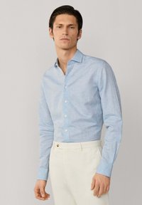 Uomo con capelli scuri che indossa una camicia azzurro chiaro con bottoni e pantaloni color avorio, in piedi davanti a uno sfondo grigio chiaro uniforme.