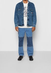 Giubbotto in denim con chiusura a bottoni, due tasche sul petto, indossato sopra una maglietta bianca con grafica. Pantaloni in denim a motivi patchwork abbinati e scarpe nere.