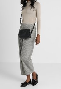 Svart läderaxelväska med rektangulär form, slät yta och minimalistisk design. Modellen bär en beige långärmad topp och rutiga byxor.