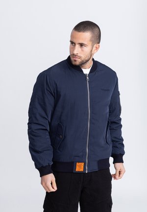 Marineblaue Bomberjacke aus glattem Stoff mit Frontreißverschluss, gerippten Bündchen und einem orangefarbenen Etikett am Saum.