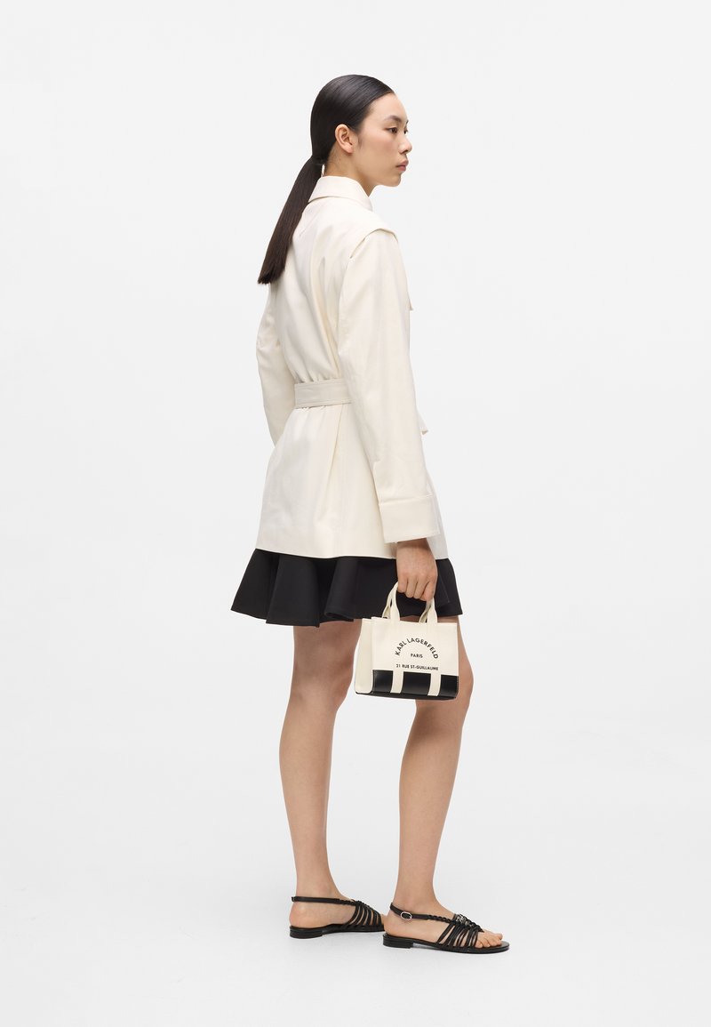 Γυναίκα με μακριά σκούρα μαλλιά, φορώντας μπεζ σακάκι με ζώνη και μαύρη φούστα, κρατώντας μια μικρή λευκή και μαύρη τσάντα, φορώντας μαύρα σανδάλια με επίπεδη σόλα.