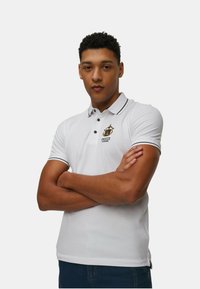 Polo shirt bianco con dettagli neri e un logo sul petto. Maniche corte, design con colletto e forma aderente. Texture morbida.