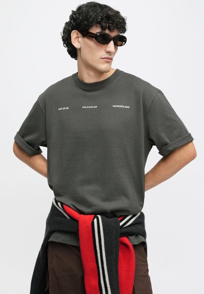 Tamno sivi majica kratkih rukava s podvrnutim manžetama, s bijelim tiskanim tekstom. Model nosi crne sunčane naočale i prugasti pleteni pulover vezan u struku.