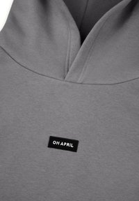 Grå hettegensere med V-hals design, laget av mykt stoff. Har en svart etikett med hvit tekst "OH APRIL" brodert på forsiden.